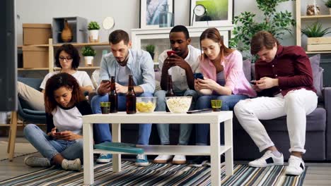 Multiracial-group-of-friends-using-smartphones-touching-screen-on-sofa-at-home