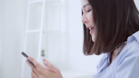 Junge-asiatische-Frau-mit-Smartphone,-während-auf-dem-Bett-nach-dem-Aufwachen-am-Morgen,-Schöne-attraktive-japanische-Mädchen-lächelnd-entspannen-im-Schlafzimmer-zu-Hause.-Spaß-im-Lifestyle-Frauen-zu-Hause-Konzept.