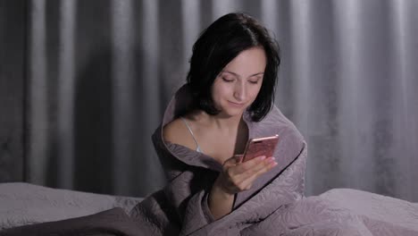 Woman-sitting-on-bed-under-blanket-and-enjoying-chatting-to-friend-on-smartphone