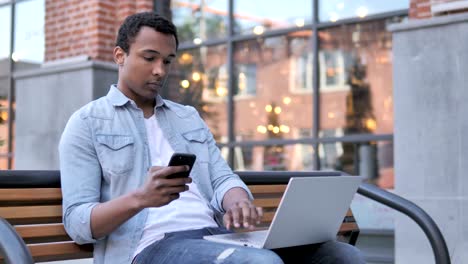 Afrikanischer-Mann-mit-Smartphone-und-Laptop,-Sitzen-auf-Bank