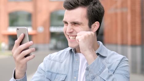 Outdoor-Young-Man-Celebrating-Success-on-Smartphone