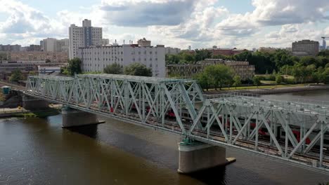 Vista-aérea-del-puente-ferroviario,-con-un-tren-en-movimiento-en-él,-a-través-del-río-que-fluye-a-través-de-una-ciudad-importante