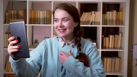 Closeup-portrait-of-young-pretty-female-student-having-a-video-call-on-the-tablet-in-the-university-library-indoors