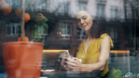 Lächelnde-junge-Frau,-die-auf-Smartphone-Bildschirm-während-schönen-Tag-im-Café