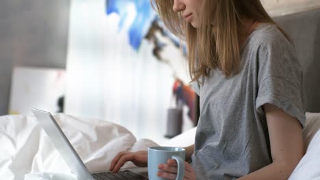 Mujer-sosteniendo-café-y-escribiendo-en-el-ordenador-portátil-en-la-cama