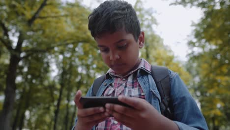 Outdoor-Porträt-afro-amerikanischen-glücklich-Schuljunge-mit-Smartphone.-Junge-Studentin,-die-auf-smobiles-Handy-schaut.-Zurück-zum-Schulkonzept.-Slow-Motion-Shot