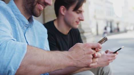 Male-Gay-Couple-Sitting-Outdoors-On-Steps-Of-Building-Looking-At-Mobile-Phones