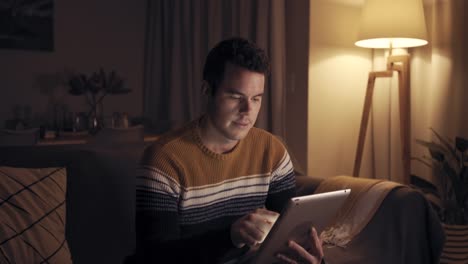 Retrato-de-un-hombre-sonriente-sentado-en-el-sofá-a-altas-horas-de-la-noche-trabajando-con-la-tableta-digital-en-casa