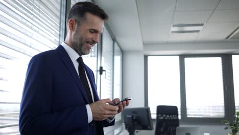 Glücklicher-Geschäftsmann-lehnt-sich-an-Bürofenster-und-tippt-auf-dem-Smartphone