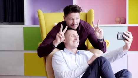 Gay-couple-using-phone-on-arm-chair-together.-Selfie-with-victory-sign.