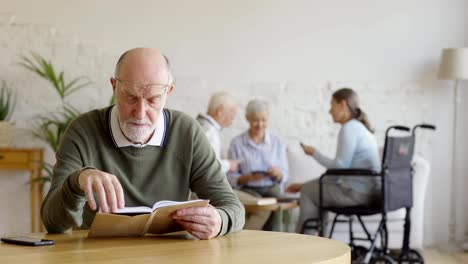 Enfoque-de-rack-de-tres-personas-mayores,-dos-mujeres-incluyendo-discapacitados-uno-y-hombre,-disfrutando-de-las-cartas-en-el-hogar-de-ancianos.-Inteligente-senior-leer-libro-sentado-en-la-mesa