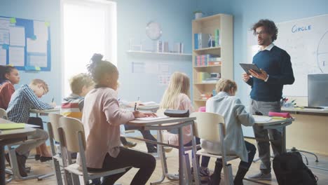 Enthusiastic-Teacher-Uses-Digital-Tablet-Computer-While-Explaining-a-Lesson-to-a-Classroom-Full-of-Diverse-Bright-Children.-In-Elementary-School-Group-of-Smart-Multiethnic-Kids-Learning