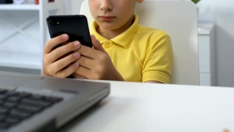 Small-boy-sitting-in-front-of-laptop,-watching-video-on-smartphone,-addiction