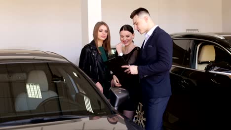 Ein-junger-lächelnder-Autoverkäufer-berät-zwei-Mädchen-im-Showroom.