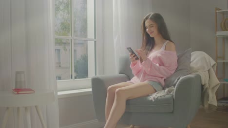 Beautiful-Young-Woman-Using-Smartphone-Smilingly-while-Sitting-on-the-Chair.-Sensual-Girl-Wearing-Cute-Pink-Sweater-and-Shorts,-Surfs-Internet,-Posts-on-Social-Media-while-Relaxing-in-Cozy-Apartment