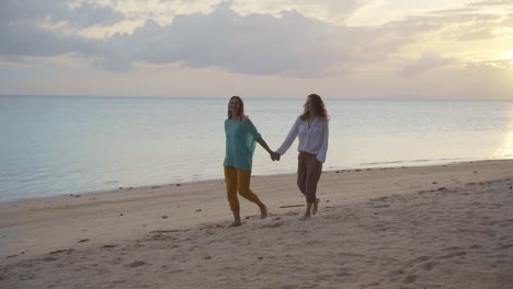 Zwei-junge-glückliche-Frauen,-die-bei-Sonnenuntergang-am-Strand-spazieren-gehen