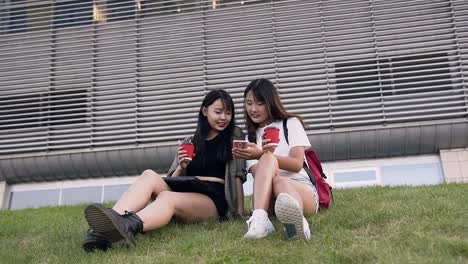 Front-view-of-charming-joyful-asian-girlfriends-which-sitting-on-the-green-grass-and-looking-at-the-phone-near-modern-building