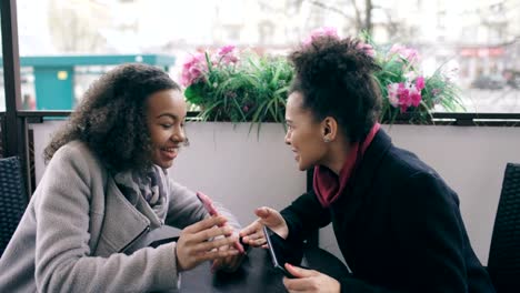 Zwei-attraktive-gemischte-Rasse-weibliche-Freunde-teilen-sich-mit-dem-Smartphone-in-Straßencafé-im-Freien