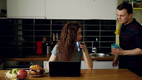 mujer-caucásica-feliz-rizada-atractiva-joven-está-sentado-en-la-cocina-trabajando-en-ordenador-portátil-beber-té,-viene-su-marido-y-ella-muestra-algo-en-laptop