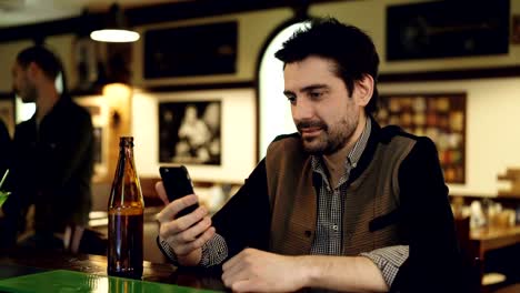 Young-male-student-is-using-smart-phone-sitting-in-fancy-bar-with-bottle-of-beer.-He-is-touching-screen-and-smiling.-Modern-ways-of-communication-concept.