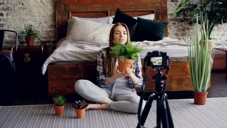 Young-smiling-blogger-in-casual-clothing-is-holding-flowers,-talking-and-recording-video-blog-for-online-vlog-about-house-plants-using-camera-on-tripod.