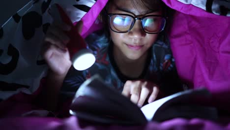 Girl-holding-flashlight-while-reading-books-blanket-at-home