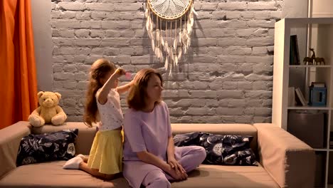 Little-daughter-brushing-her-mother-hair-with-hairbrush,-sitting-on-sofa-in-modern-living-room,-family-concept-indoors