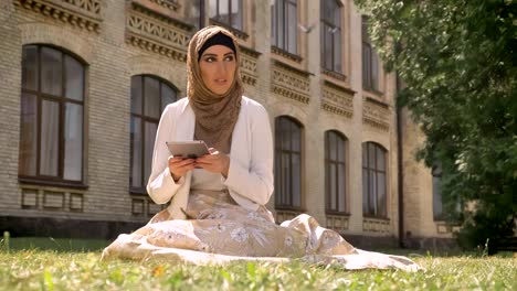 Beautiful-muslim-woman-in-hijab-typing-on-tablet-and-sitting-on-grass-in-park-near-building,-charming-female-with-gadget