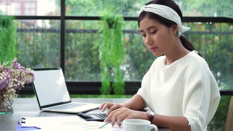 Beautiful-young-smiling-asian-woman-working-on-laptop-while-sitting-in-living-room-at-home.-Asian-business-woman-working-document-finance-and-calculator-in-her-home-office.-Enjoying-time-at-home.