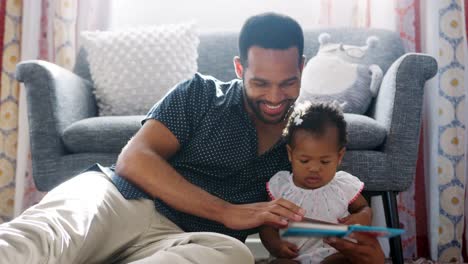 Father-sitting-on-floor-at-home-with-baby-daughter-reading-book-together---shot-in-slow-motion