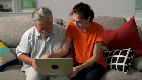 Tochter-Unterricht-ihres-Vaters-Computerkenntnisse-im-Wohnzimmer.-Asiatischer-Mann-mit-weißem-Bart-und-junge-Frau-sitzt-im-Wohnzimmer-mit-Laptop.-Familie-Senior-Lifestyle-Konzept.