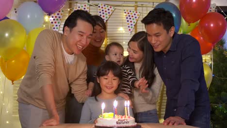 A-cute-little-girl-sitting-at-table-and-blowing-candles-on-birthday-cake-while-her-family-standing-behind-and-sing-a-song-to-her.-She-gives-a-big-smile-and-her-family-applaud-her.-In-slow-motion.