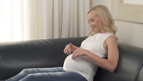 A-side-view-of-a-pretty-pregnant-blonde-is-sitting-on-the-sofa-and-stroking-her-belly,-she-is-looking-at-it-and-smiling