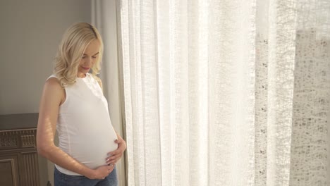 Ein-seitlicher-Blick-auf-eine-schöne-Blonde-am-Fenster-stehen-und-streichelte-ihren-Bauch.-Sie-befasst-sich-mit-der-Kamera-und-lächelt,-dann-schaut-auf-den-Bauch-mit-Zärtlichkeit