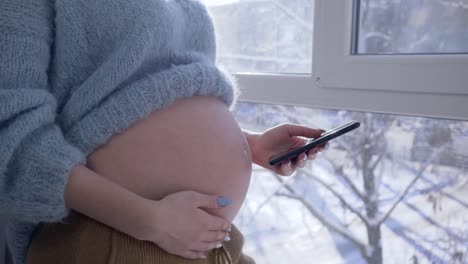 niña-embarazada-utiliza-teléfono-celular-y-acaricia-su-estómago-desnudo-parado-por-una-gran-ventana-en-el-apartamento