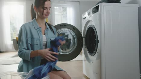 Beautiful-Smiling-Brunette-Young-Woman-Sits-in-Front-of-a-Washing-Machine-in-Homely-Jeans-Clothes.-She-Loads-the-Washer-with-Dirty-Laundry.-Bright-and-Spacious-Living-Room-with-Modern-Interior.