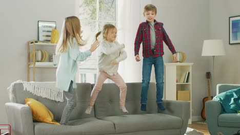 Zwei-niedliche-kleine-Mädchen-und-junge,-bezaubernde-haben-Spaß,-springen-hoch-auf-einer-Couch-zu-Hause.-Glückliche-Kinder-spielen-im-sonnigen-Wohnzimmer.-In-Zeitlupe.