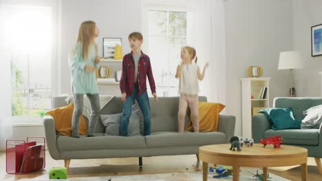 Two-Cute-Little-Girls-and-Young-Adorable-Boy-Have-Fun,-Jumping-High-on-a-Couch-at-Home.-Happy-Kids-Dancing-on-a-Sofa-in-the-Sunny-Living-Room.