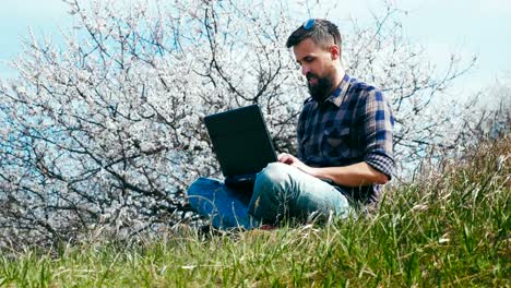 Plano-de-plataforma-rodante.-Hombre-con-una-barba-utiliza-portátil-en-la-naturaleza-cerca-de-flor-árbol