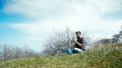 Hombre-con-una-barba-utiliza-una-tableta-en-naturaleza-cerca-de-flor-árbol