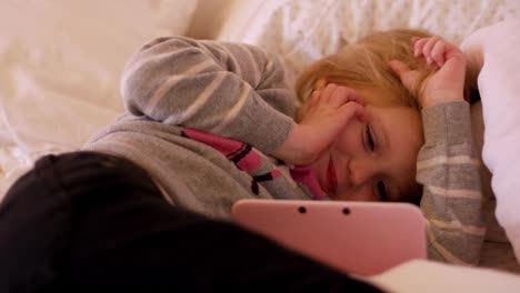 A-little-girl-laying-on-her-bed-watching-a-show-on-her-handheld-gaming-system,-and-she-smiles-and-waves