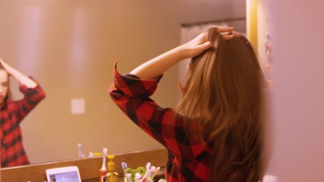 A-young-girl-doing-her-hair-in-the-mirror-in-the-bathroom,-and-having-a-video-chat-on-her-tablet