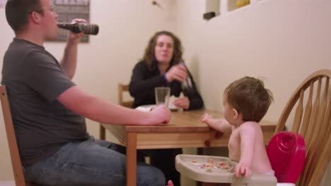 Una-familia-cenando-juntos-en-la-mesa-de-la-cocina,-con-un-niño-pequeño-sentado-en-un-asiento-de-silla-alta