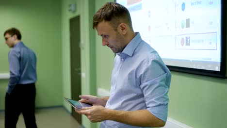 Young-man-uses-tablet-to-a-large-audience.-Image-from-the-projector-on-the-background.-Digress-from-working-on-the-tablet-to-see-who-sitting-in-the-audience