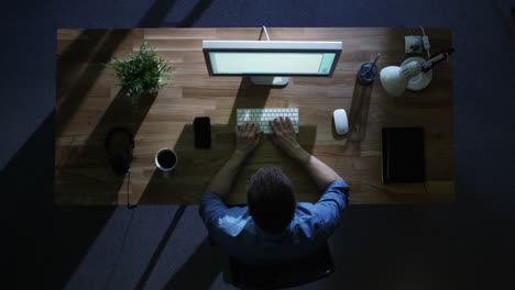 Vista-superior-del-programador-hombre-trabajando-en-su-computadora-de-escritorio-en-la-noche.-Su-mesa-está-iluminada-por-frío-azul-luz-desde-fuera.