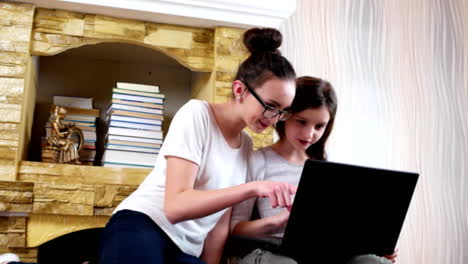 chica-con-gafas-mirando-a-su-hermana-pequeña-que-hacer-sus-deberes-en-el-ordenador-portátil,-adolescentes-sentados-juntos