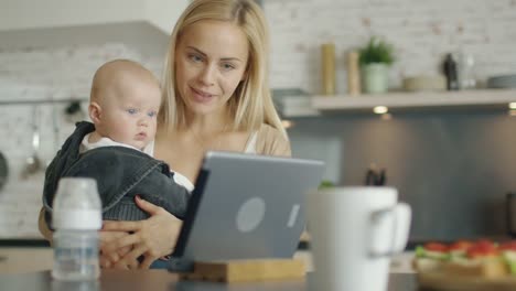 Die-Küche-hält-die-Mutter-ihr-Baby.-Beide-schauen-Sie-sich-etwas-Interessantes-auf-dem-Bildschirm-eines-Tablet-Computers.