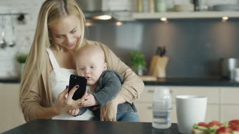 La-joven-madre-sostiene-a-su-bebé-mientras-está-en-la-cocina.-Ella-también-utiliza-smartphone-con-el-que-cute-Baby-interactúa.