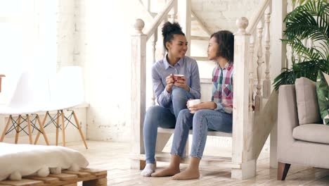 Two-african-american-curly-girls-sistres-sitting-on-stairs-have-fun-laughing-and-chatting-together-at-home