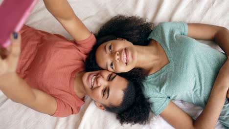 Top-view-of-cheerful-mixed-race-funny-girls-making-selfie-portrait-on-bed-in-bedroom-at-home